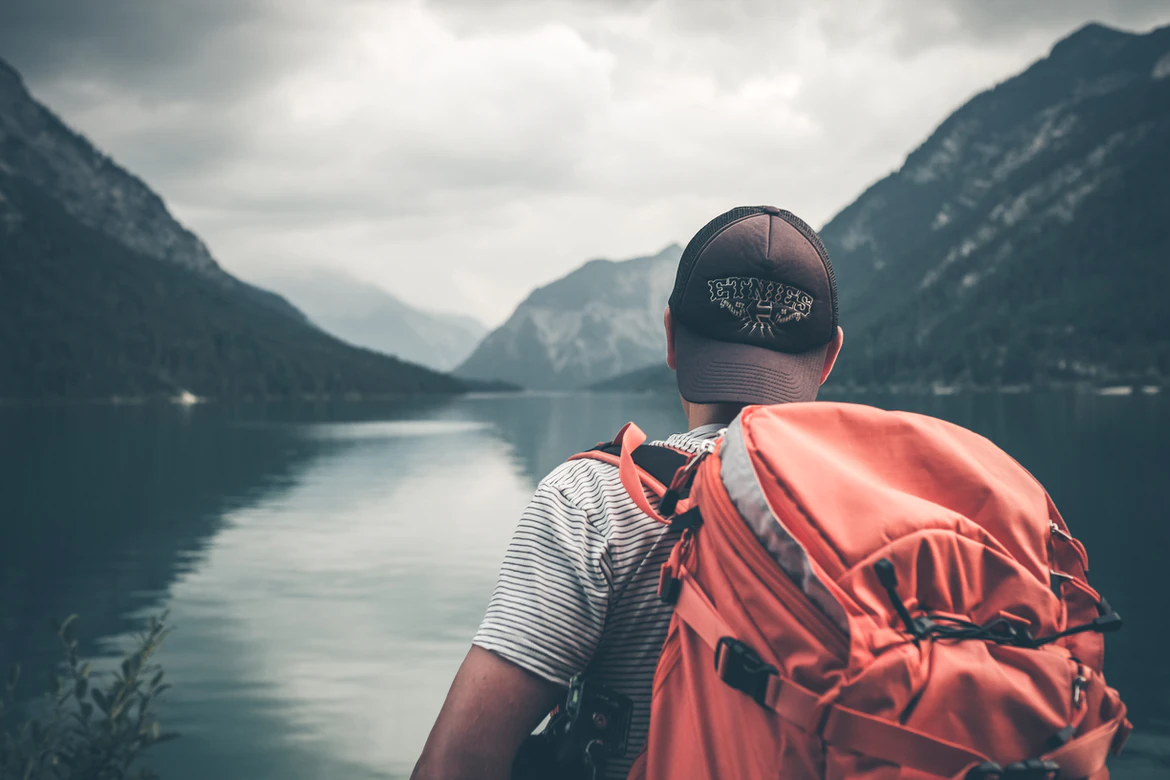 A traveller with a big orange backpack looks out over a lake and mountains.